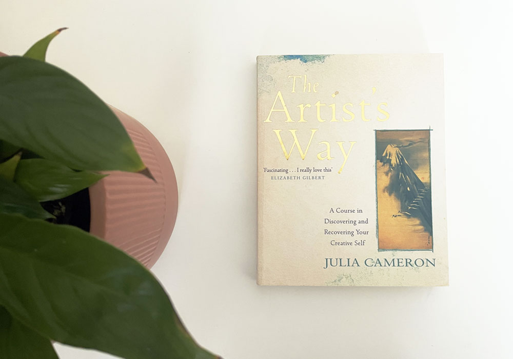 The Artists Way book by Julia Cameron on a white table with a plant on the left